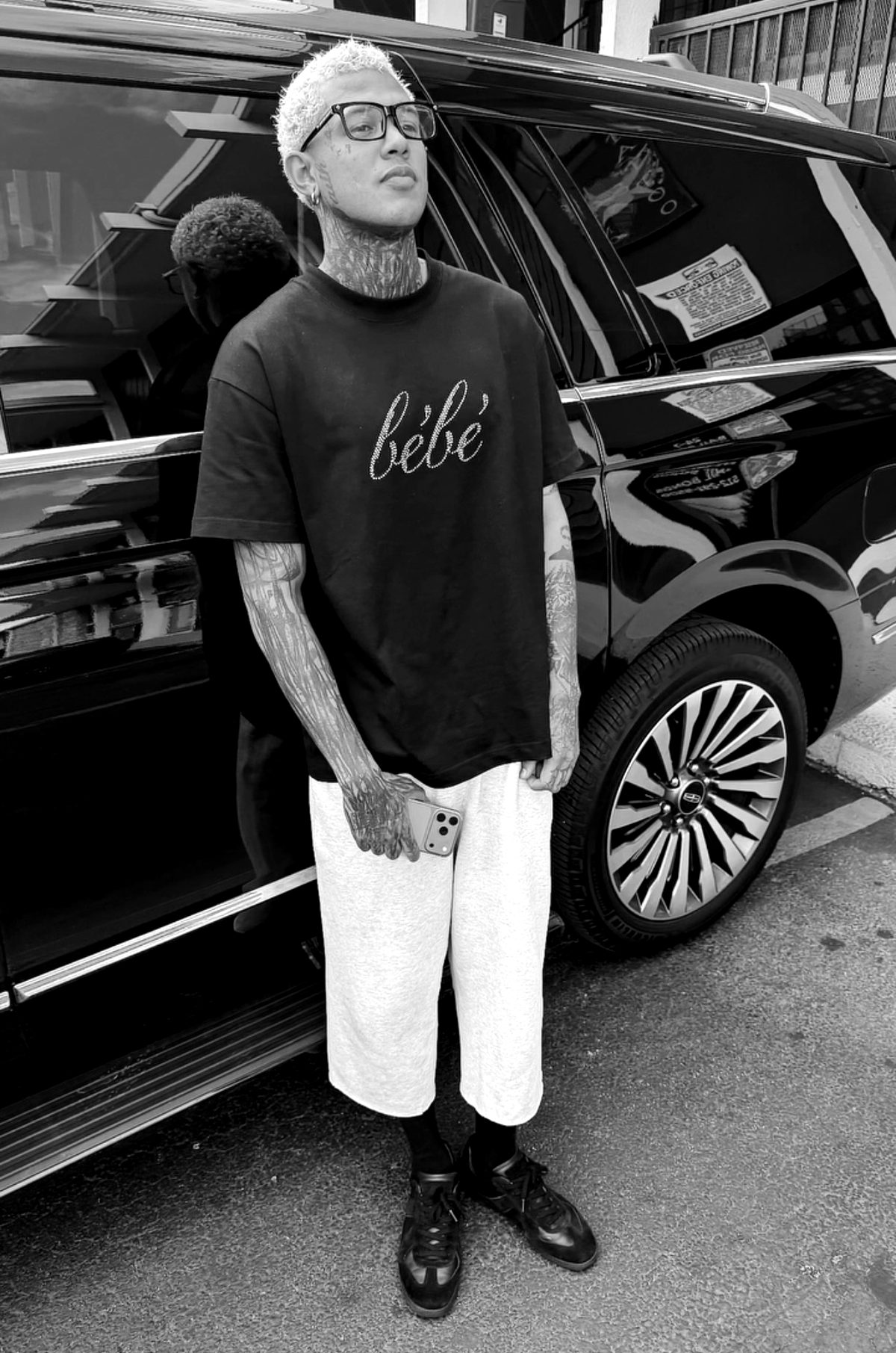 Seven Avanti standing beside a black SUV, wearing glasses and a black tee, tattooed arms and neck